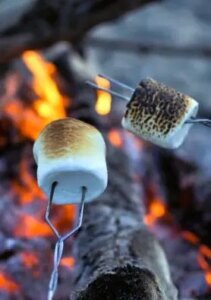 Fotografia em close-up das mãos de um casal segurando dois espetos de madeira com marshmallows queimando sobre uma fogueira de chão feita de tijolos cinzas, à noite, na grama. O marshmallow de cima está em chamas. É possível ver uma aliança dourada no dedo anelar da mão masculina que segura o espeto de baixo e um anel delicado na mão feminina. O fundo é escuro.