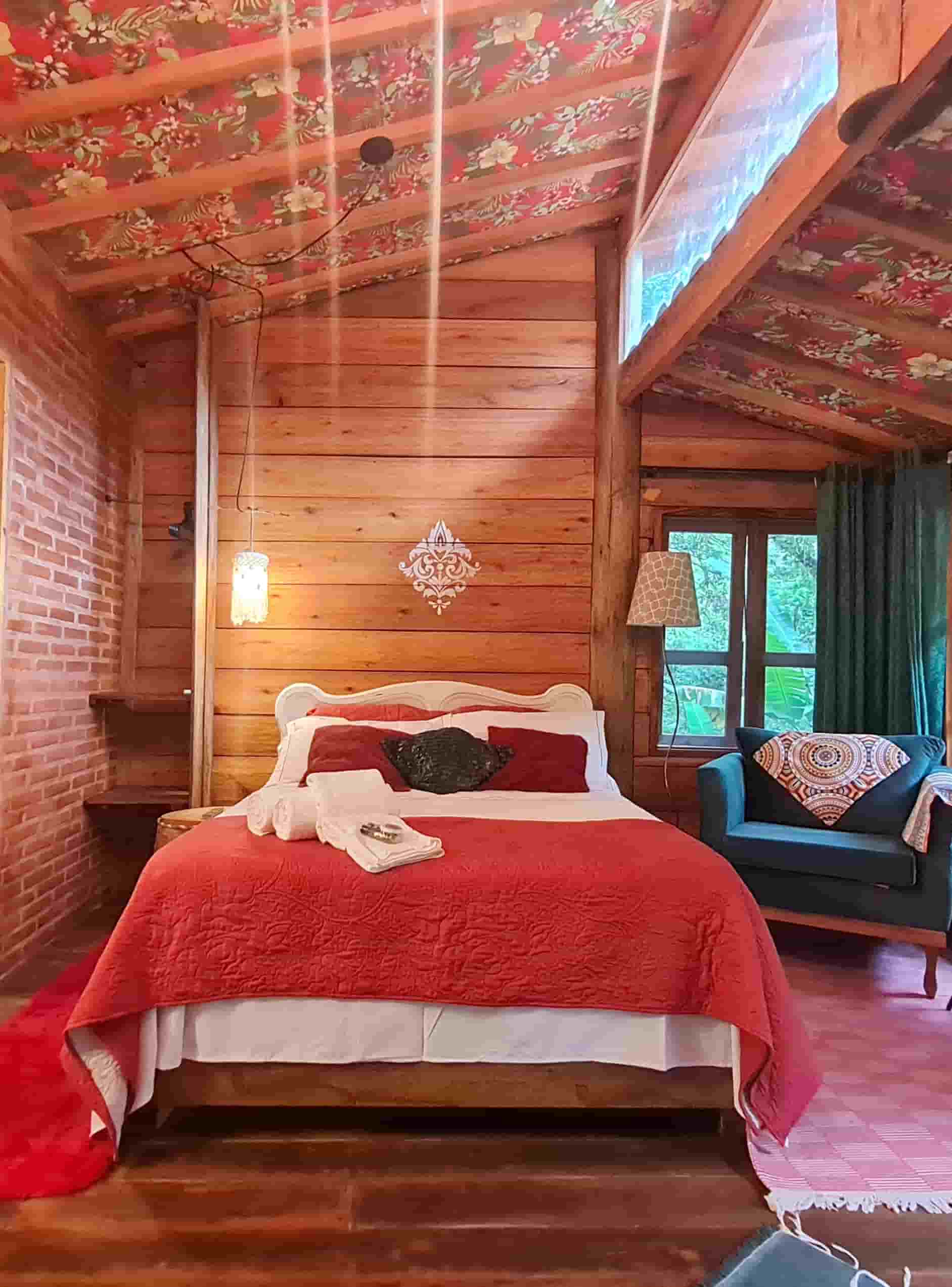 Quarto aconchegante da Cabana Kel com forro de chita artesanal, manta vermelha e janelas de madeira voltadas para a mata em Cunha.