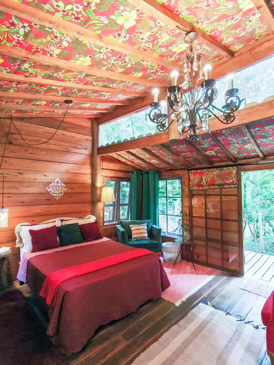 Quarto aconchegante da Cabana Kel com forro de chita artesanal, manta vermelha e janelas de madeira voltadas para a mata em Cunha.